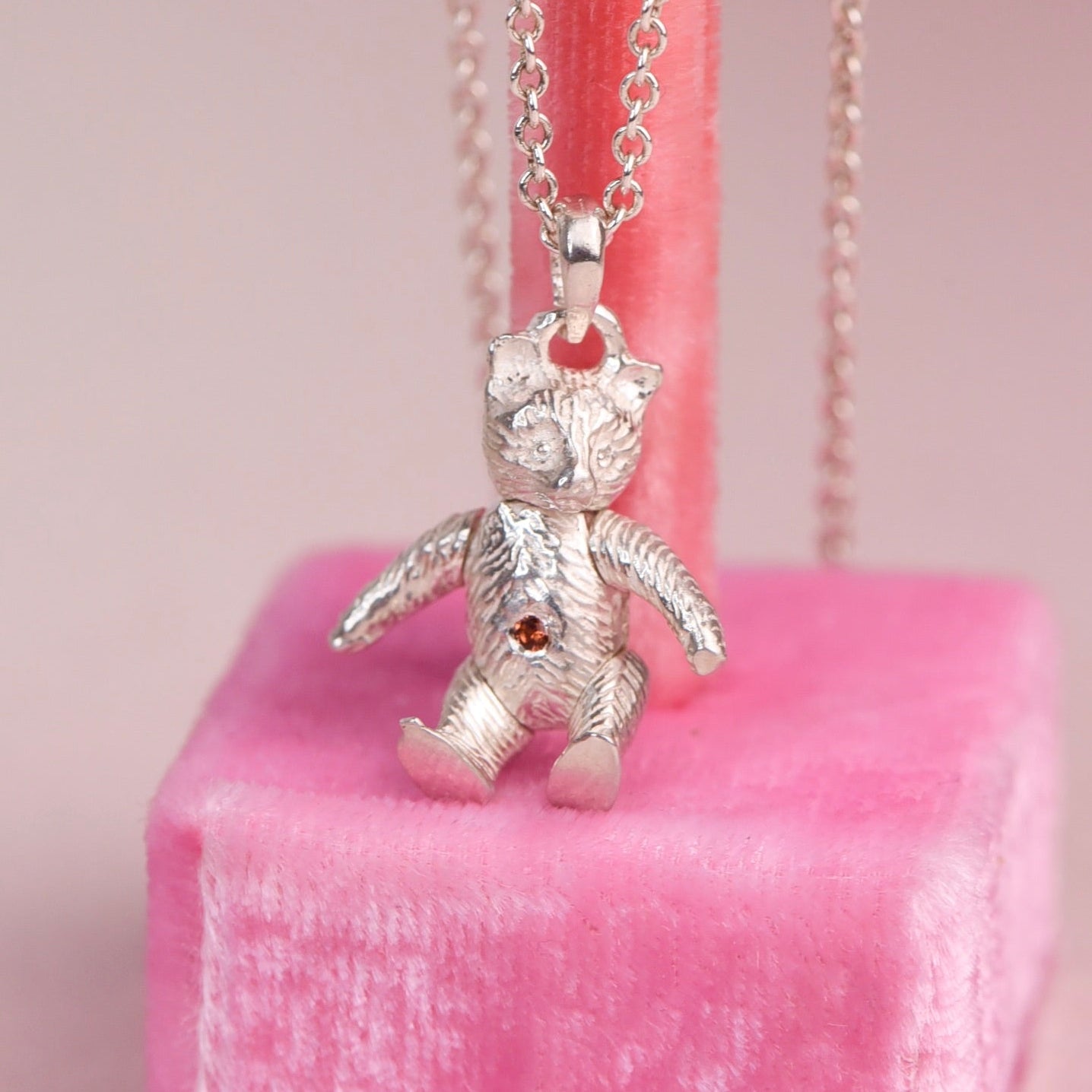 A silver teddy bear pendant necklace with a sorange diamond on belly. displayed on a pink velvet stand against a soft pink background.