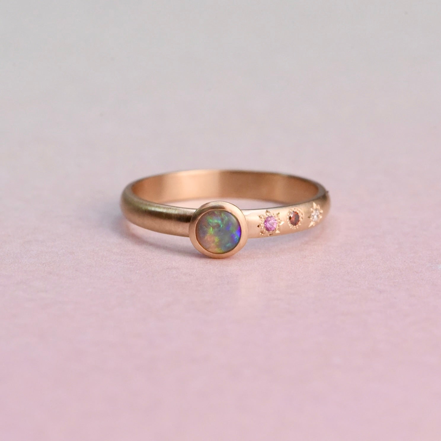 Front view of the Asymmetrical Black Opal and Sapphire 14k Gold Ring by Corkie Bolton Jewelry, featuring a Lightning Ridge black opal bezel-set in recycled gold with pink, red, and white stones in hand-engraved star settings.
