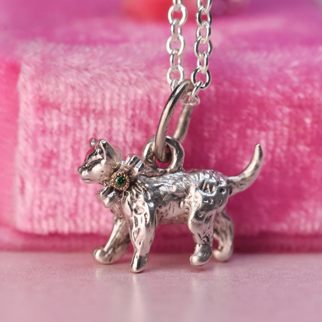 Close up of a vintage Beaucraft sterling silver kitten charm featuring a green diamond set in a 14k yellow gold bow. with a pink velvet background.