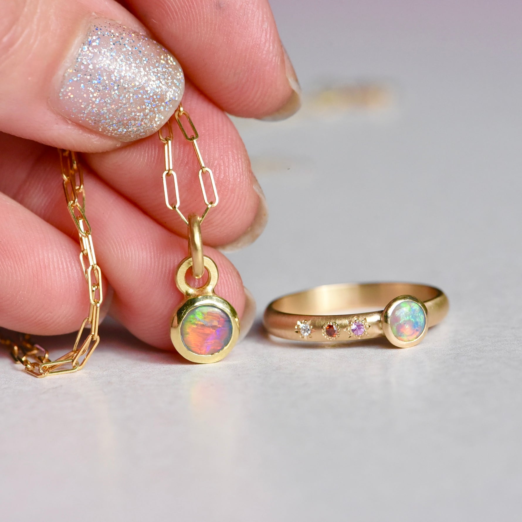 Gold necklace and ring with opal stones held by a hand with glittery nail polish.