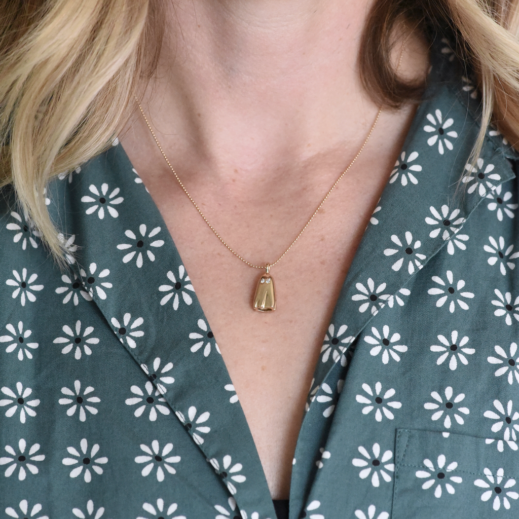 Solid 10k yellow gold ghost charm with diamond eyes on a model's neck wearing a floral shirt, by Corkie Bolton Jewelry.
