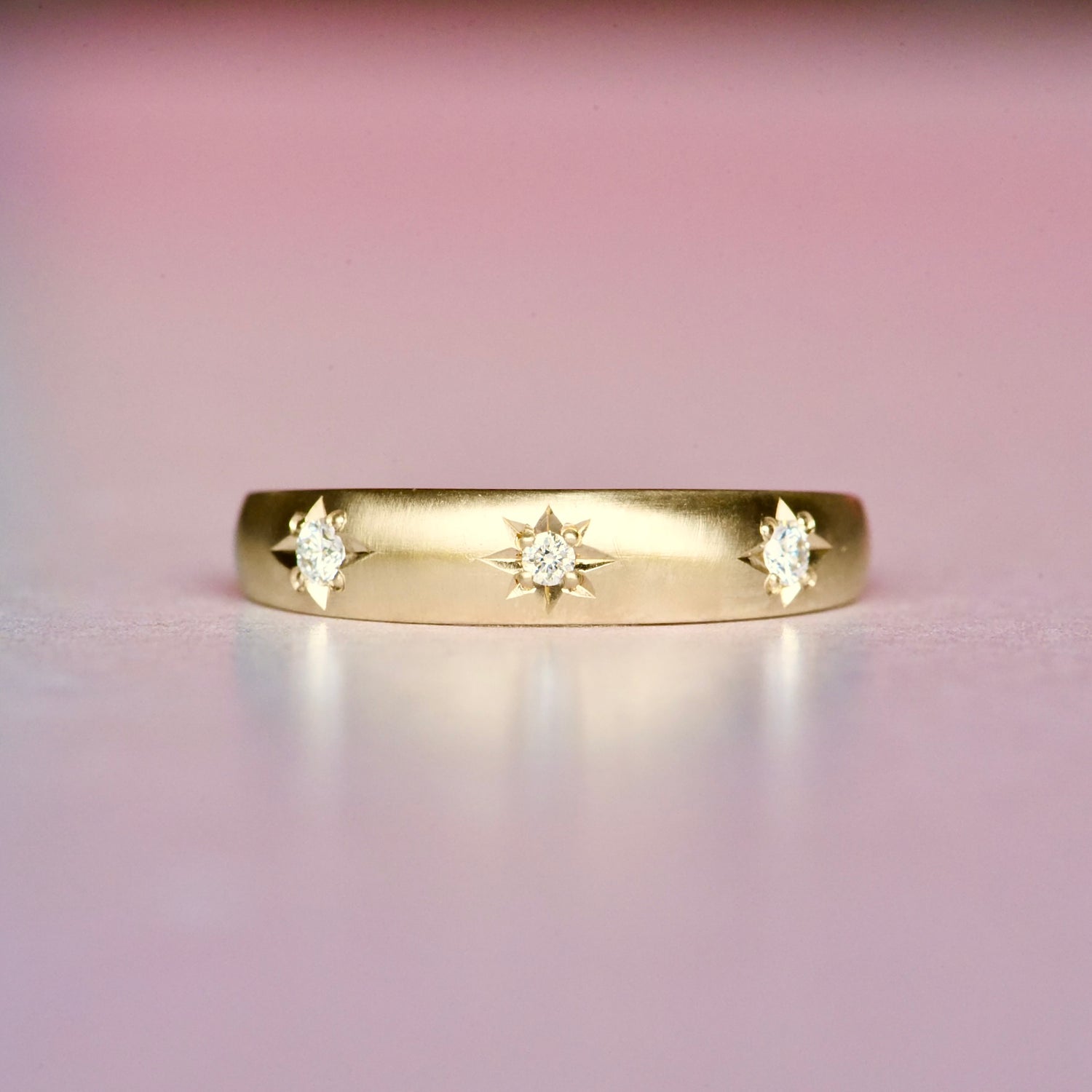 Tapered 10k gold ring with three diamonds in star settings, front view on a soft pink background