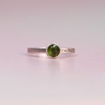 Sterling silver bezel ring with a rosecut green tourmaline and “One Day At A Time” engraved band, photographed from the front on a soft pink background.