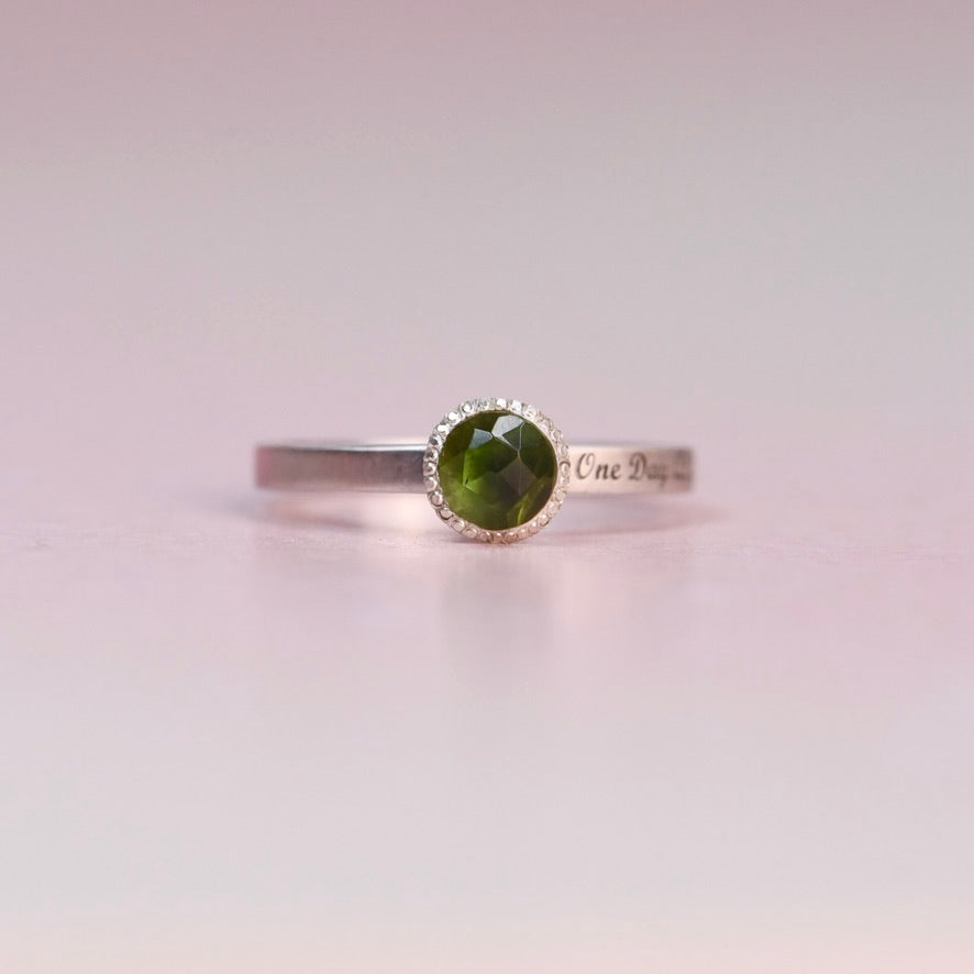 Sterling silver bezel ring with a rosecut green tourmaline and “One Day At A Time” engraved band, photographed from the front on a soft pink background.