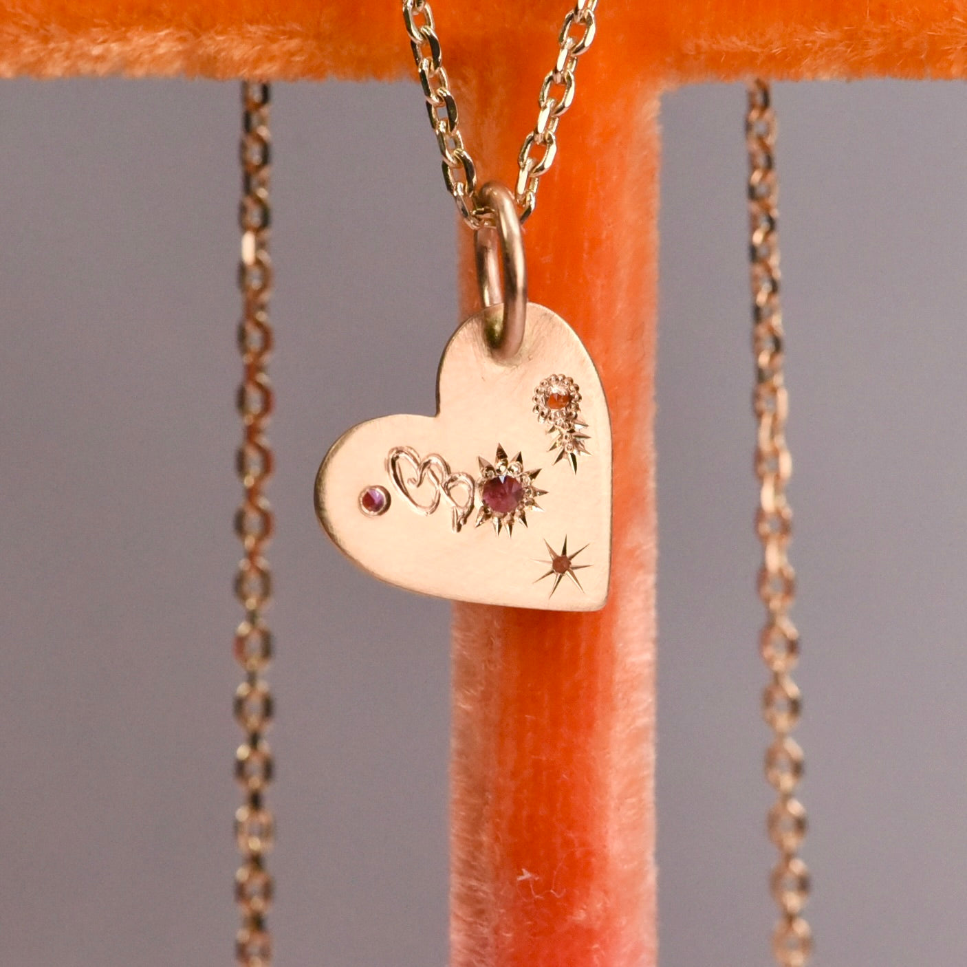 Back of Misfit Heart Pendant engraved with a tiny flower and stars, showing fine hand engraving in 14k gold. Orange velvet stand.