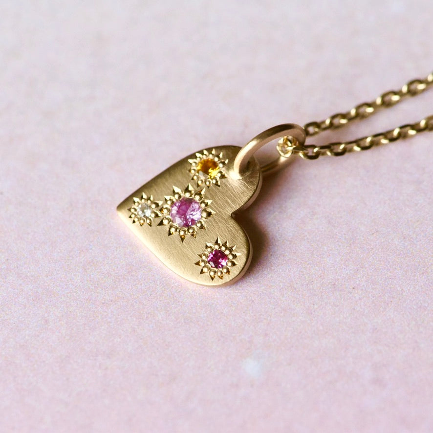 Top view of the Misfit Heart Pendant showing asymmetrical star-set sapphires and diamond with a warm yellow 10k chain. On a pink background.