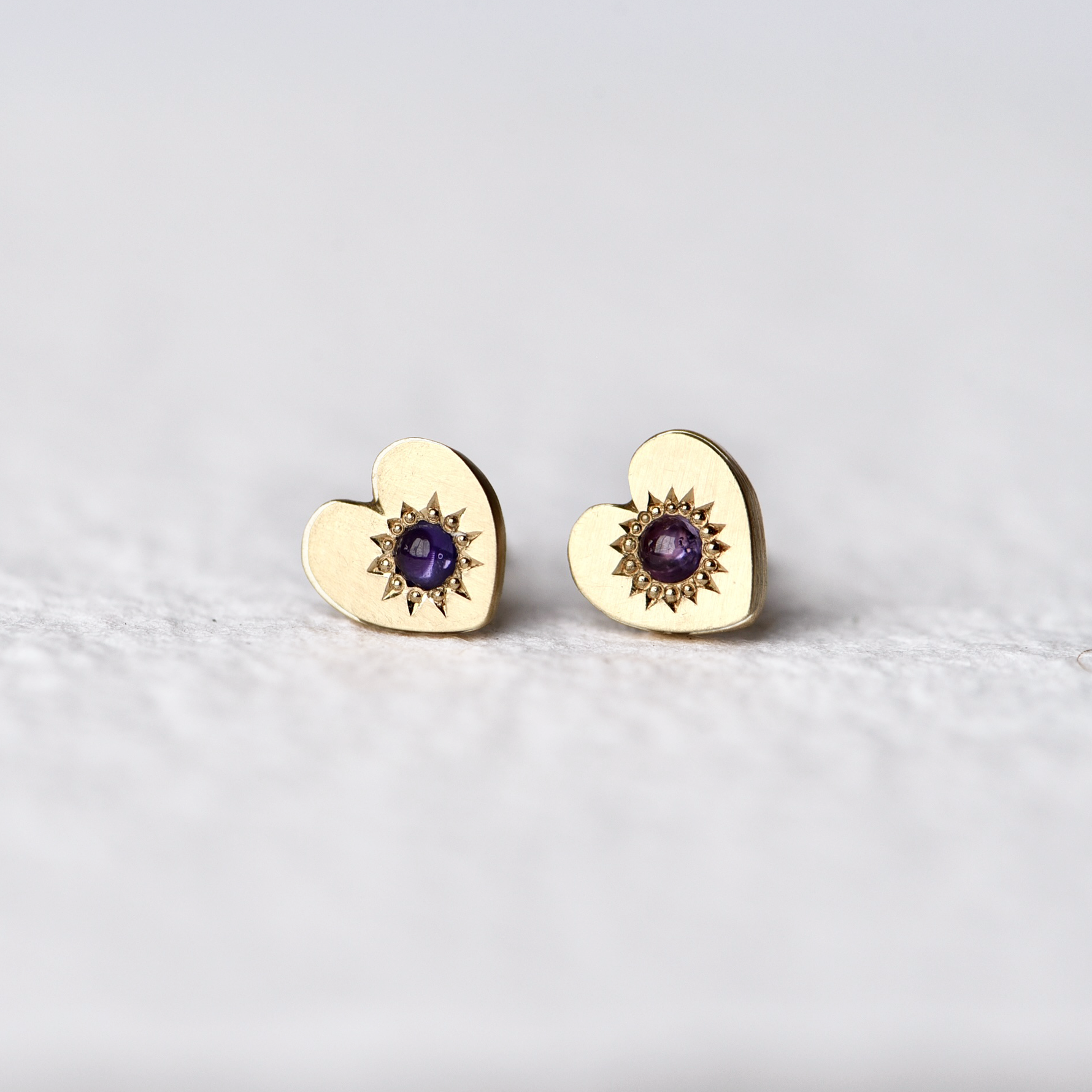 Close-up of Misfit 14k Gold Studs on a white background: A detailed view of the Misfit 14k gold heart-shaped studs, each featuring a color-treated purple sapphire cabochon, showcasing their unique handcrafted design and matte finish.