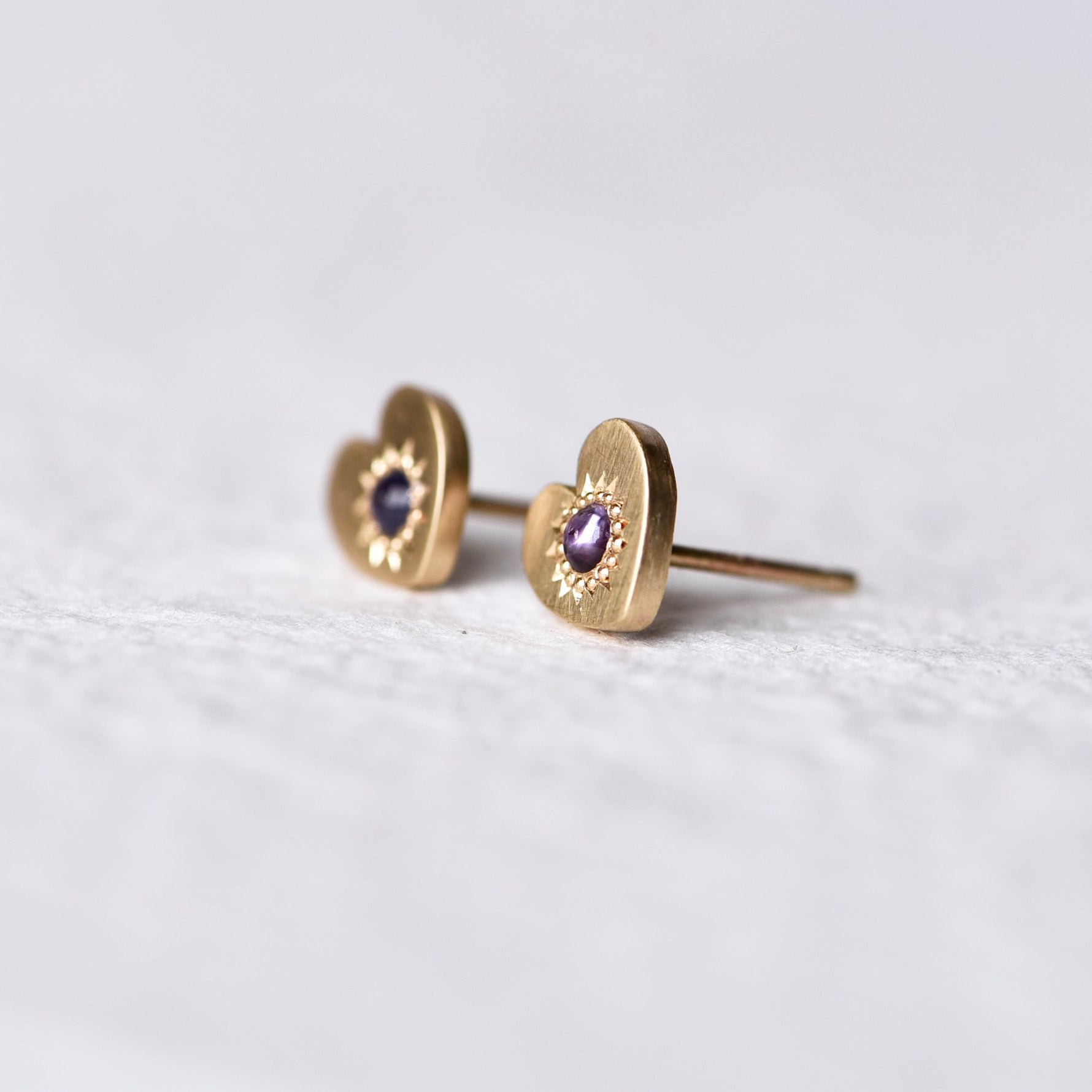 Side angle of Misfit 14k Gold Studs on a white background: A detailed view of the Misfit 14k gold heart-shaped studs, each featuring a color-treated purple sapphire cabochon, showcasing their unique handcrafted design and matte finish.