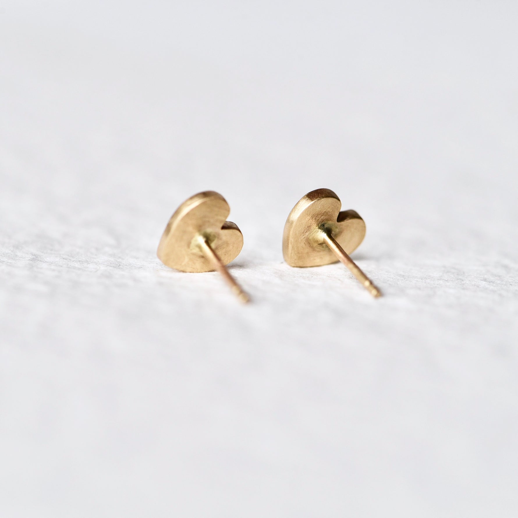 Back side of Misfit 14k Gold Studs on a white background: A detailed view of the Misfit 14k gold heart-shaped studs, each featuring a color-treated purple sapphire cabochon, showcasing their unique handcrafted design and matte finish.