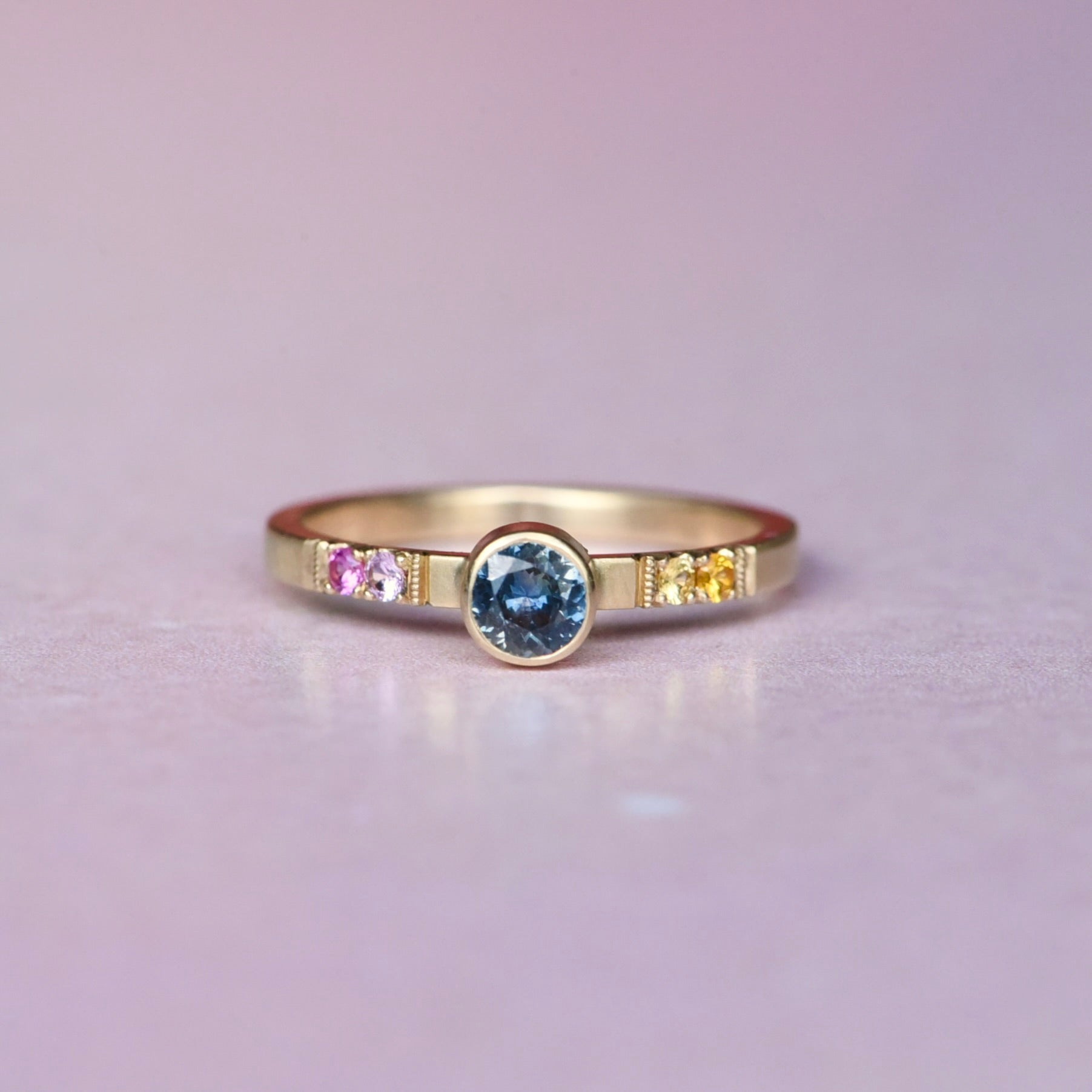 Front view of a one-of-a-kind Montana sapphire ring in 10k yellow gold, featuring a .50ct hand-selected center stone and an ombré of orange, yellow, and pink sapphires in castle settings with milgrain detail.