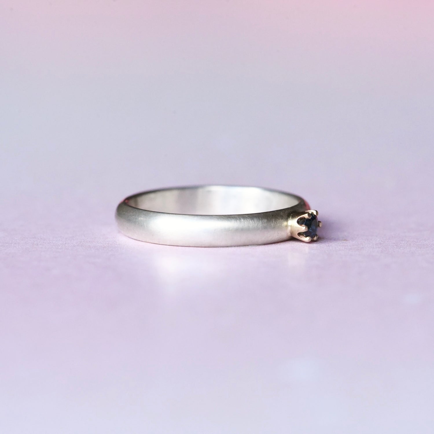 Side angle of a silver ring with a 14k yellow gold 6 prong setting with a blue faceted sapphire. The ring is on a light purple background