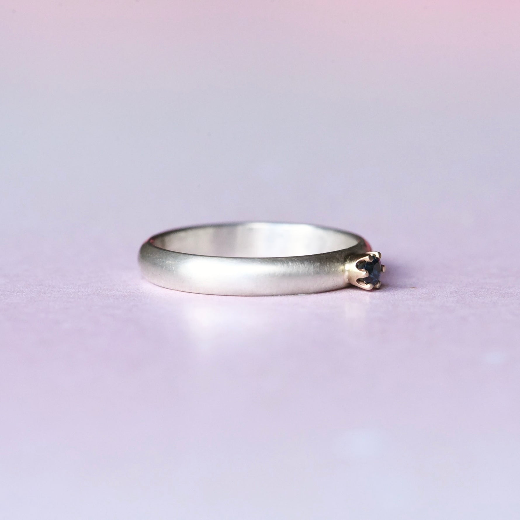 Side angle of a silver ring with a 14k yellow gold 6 prong setting with a blue faceted sapphire. The ring is on a light purple background