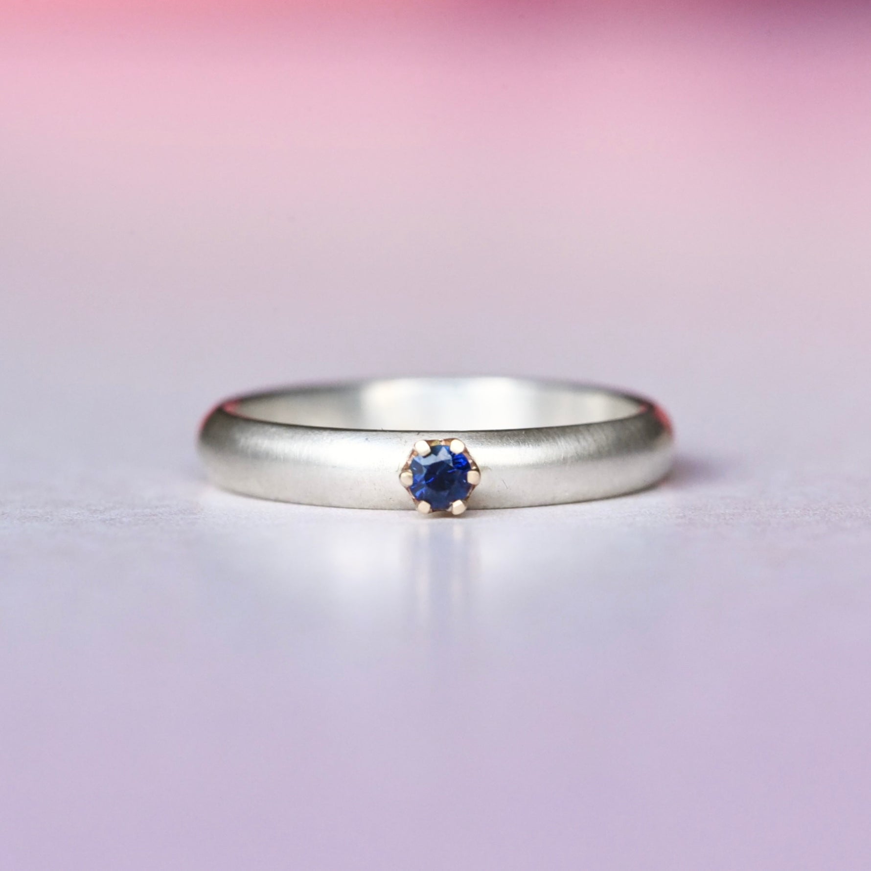 Silver ring with a 14k yellow gold prong set blue sapphire on a gradient pink and purple background