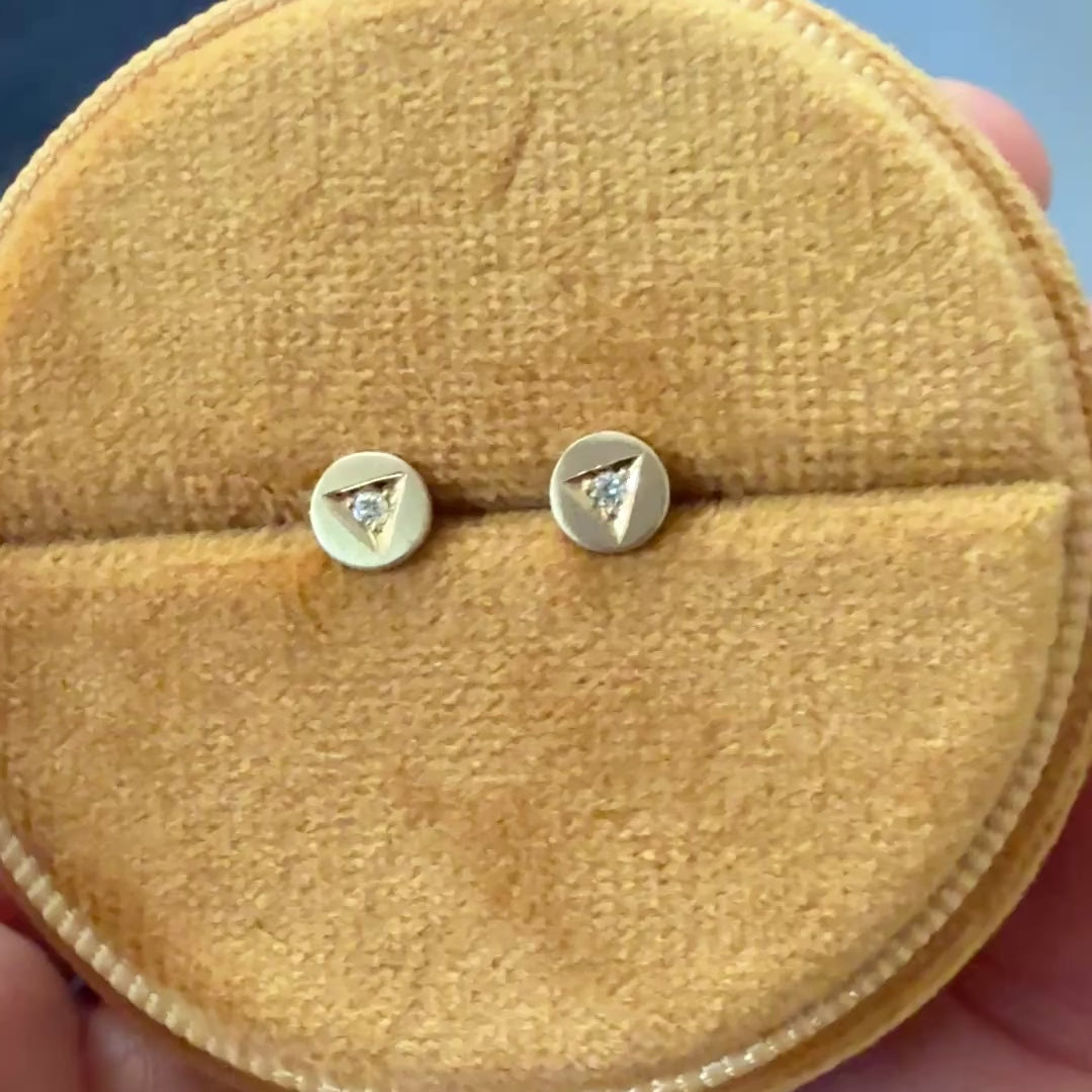 Video of Handcrafted 14k gold diamond stud earrings by Corkie Bolton Jewelry featuring unique triangle engravings. These artisan-made earrings highlight delicate, hand-engraved details with a sparkling diamond centerpiece in each stud, blending minimalism with fine craftsmanship for a timeless design. they are in a display velvet box that is round.