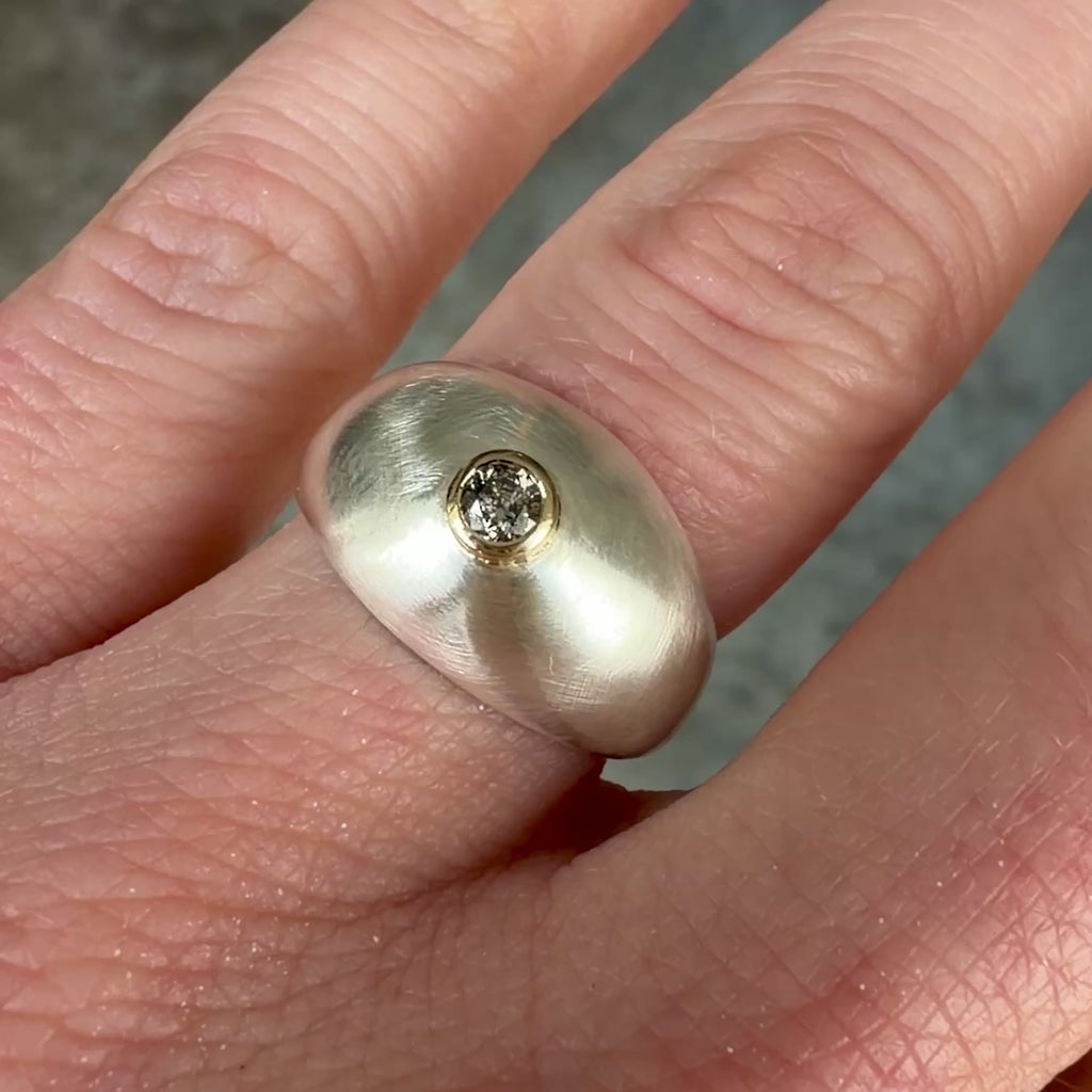 A close-up video still of a chunky brushed sterling silver dome ring with a 14k yellow gold bezel set with a faceted salt and pepper diamond, worn on a finger.