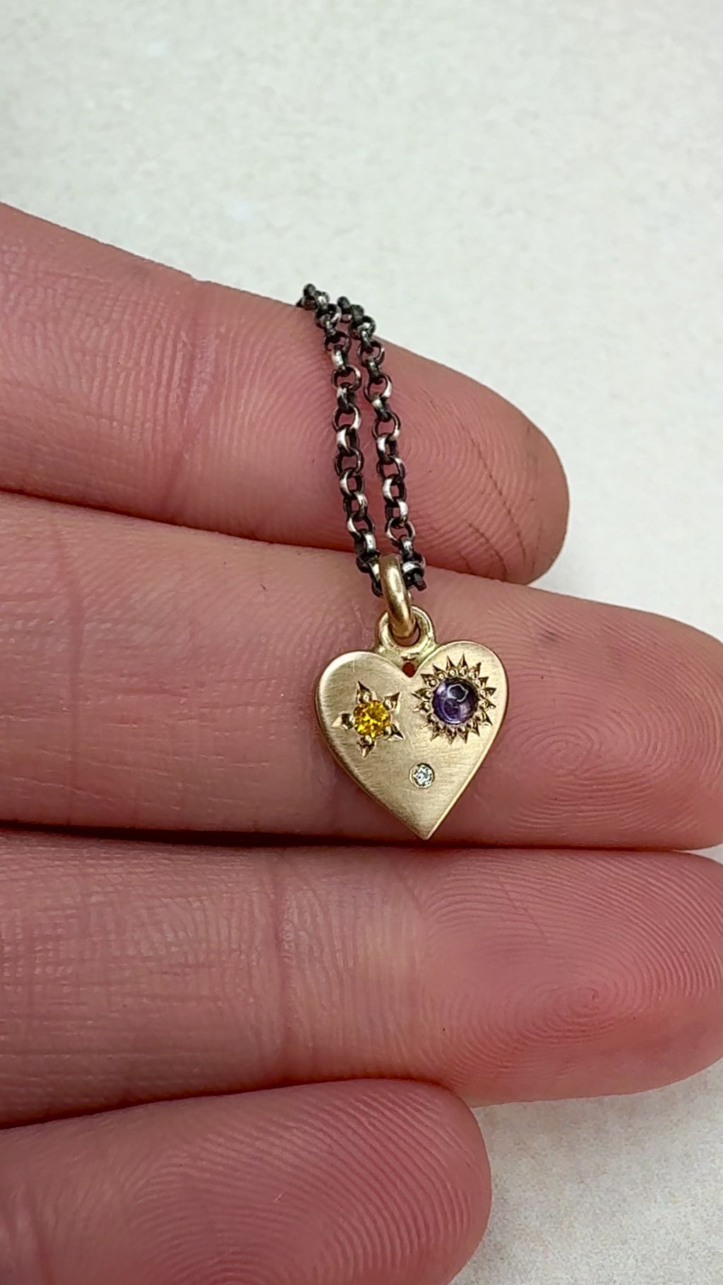 Video of the HELL YES 14k Gold Sapphire and Diamond Misfit Heart Necklace rotating under natural light, showing the yellow and purple sapphires, diamond sparkle, and hand-engraved “HELL YES” on the back. Handmade by Corkie Bolton Jewelry.