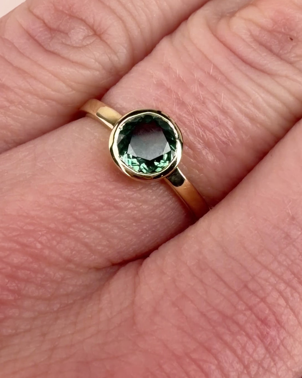 Video of a teal sapphire gold ring turning on a finger, showing the stone in a 14k yellow gold bezel