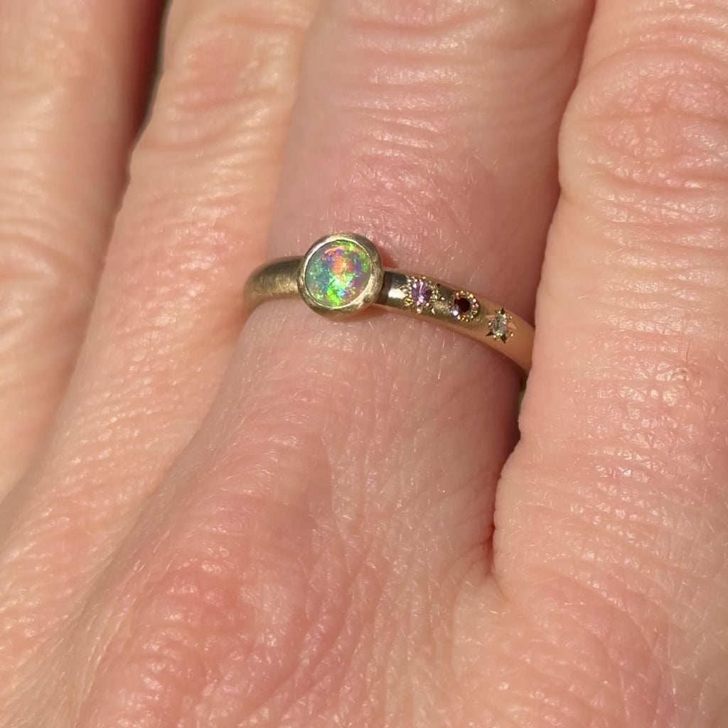 ideo of the Asymmetrical Black Opal + Sapphire 14k Gold Ring by Corkie Bolton Jewelry, capturing the opal’s vivid play of color with flashes of green, blue, and neon orange as the light moves.