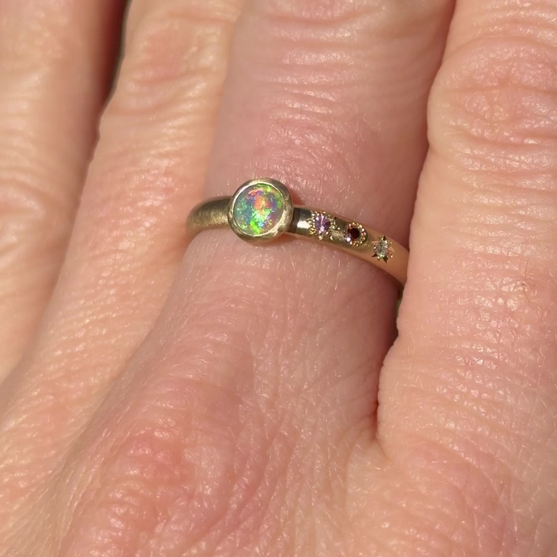 ideo of the Asymmetrical Black Opal + Sapphire 14k Gold Ring by Corkie Bolton Jewelry, capturing the opal’s vivid play of color with flashes of green, blue, and neon orange as the light moves.