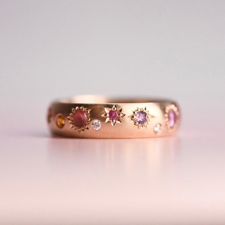 Mid-century 10k gold band reimagined with hand-engraved star settings and colorful pastel sapphires and diamonds, photographed on a soft pink background.