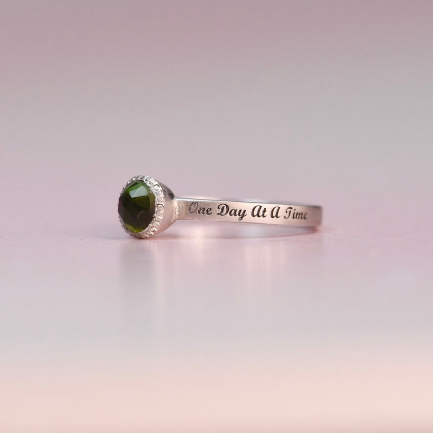 Silver ring with green gemstone and 'One Day At A Time' engraving on a light pink background