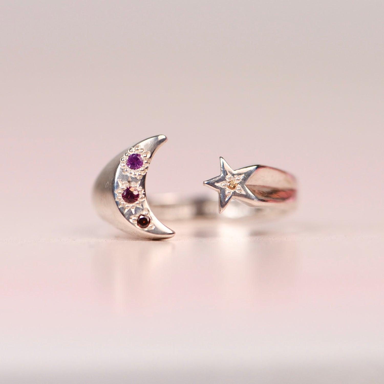 Vintage sterling silver open ring with crescent moon set with hand engraved purple pink and orange sapphires and orange diamond, and a star set with a yellow sapphire, displayed on a neutral surface by Corkie Bolton Jewelry