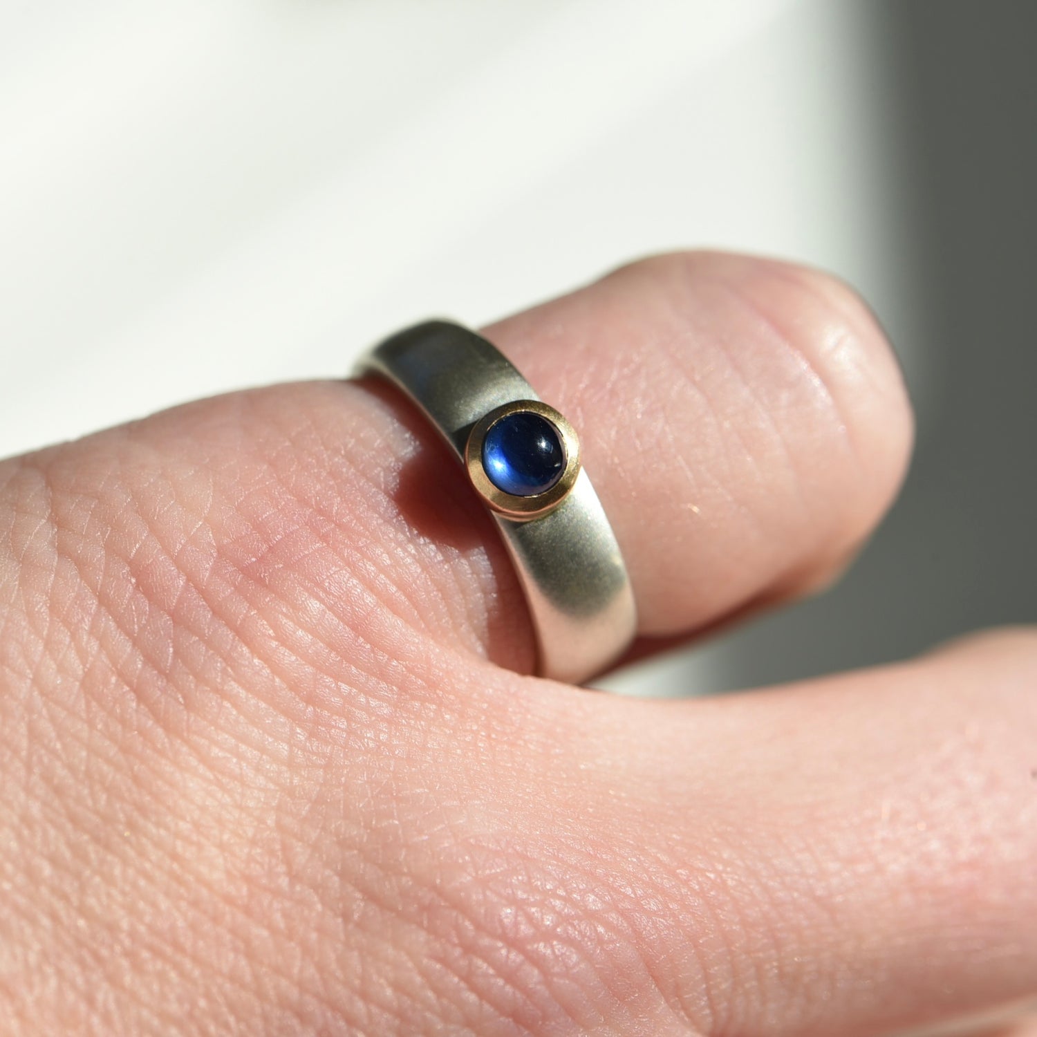 This image is of a sterling silver wide ring that has a 14k gold bezel featuring a blue sapphire cabochon. Created by Corkie Bolton.