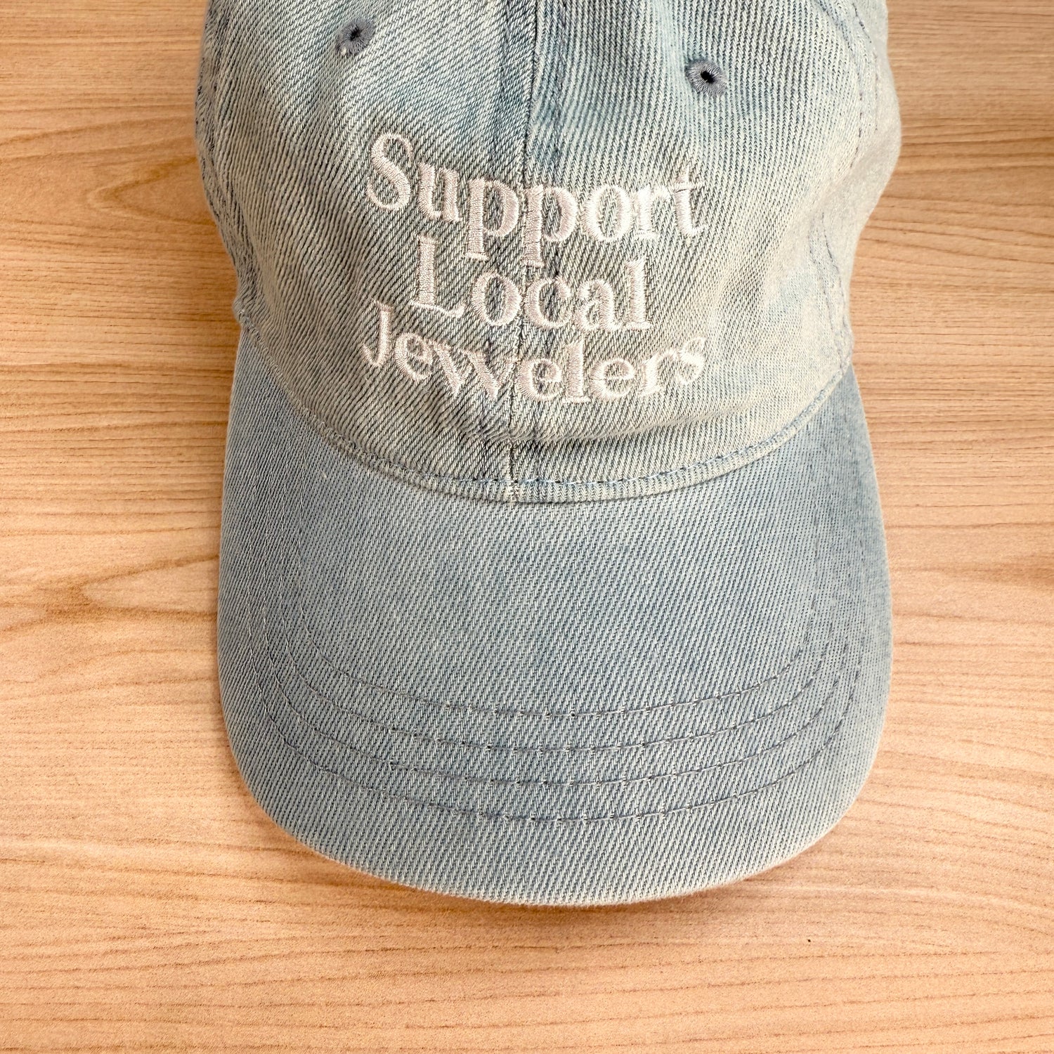 Close-up detail of the white Support Local Jewelers embroidery on the light denim cotton hat, showing the clean stitching and textured 6-panel fabric. Designed by Corkie Bolton Jewelry to celebrate independent makers.