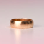 A plain polished 14k yellow gold wide band vintage ring displayed on a soft blush pink surface, before being transformed into a Reborn Vintage piece.