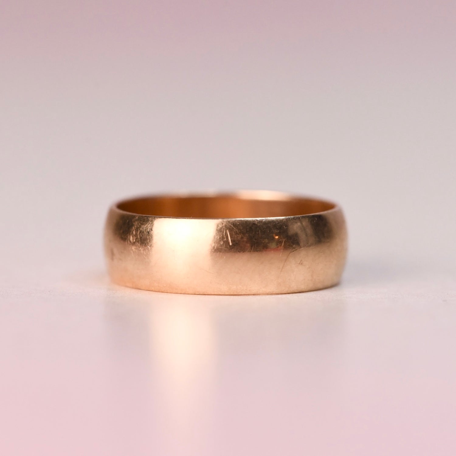 A plain polished 14k yellow gold wide band vintage ring displayed on a soft blush pink surface, before being transformed into a Reborn Vintage piece.