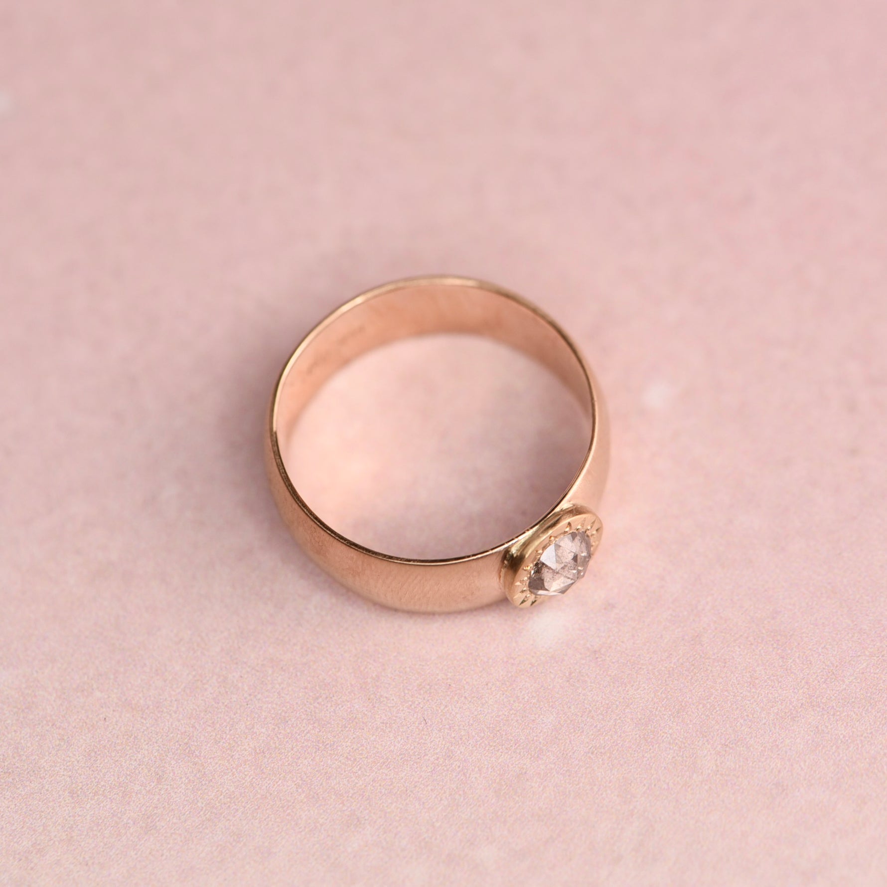 Angled view of a wide 14k yellow gold vintage band ring with an 18k yellow gold hand-engraved bezel set with a 0.40ct salt and pepper rose cut diamond, displayed on a soft blush pink surface.