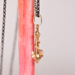 Side view of a vintage mid-century 18k yellow gold anchor and rope charm pendant with star set white diamond hanging on an oxidized black rolo chain against a pink display stand, showing sculptural rope detail and 14k gold jump ring connector, by Corkie Bolton Jewelry