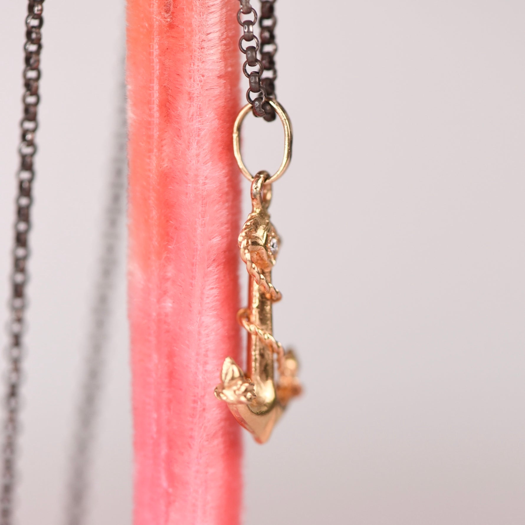 Side view of a vintage mid-century 18k yellow gold anchor and rope charm pendant with star set white diamond hanging on an oxidized black rolo chain against a pink display stand, showing sculptural rope detail and 14k gold jump ring connector, by Corkie Bolton Jewelry