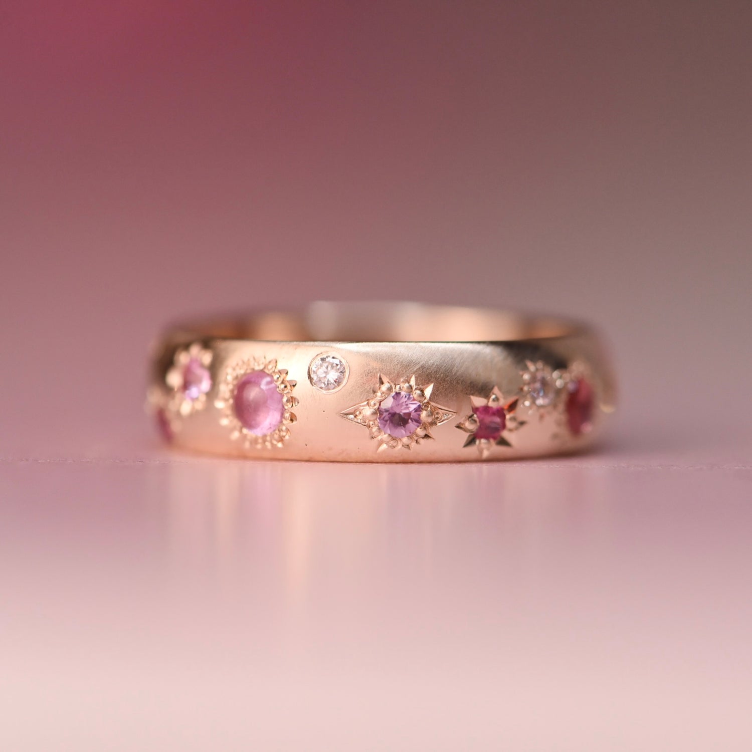 Gold ring with pink gemstones on a pink background. Close-up of the Vintage Confetti Diamond + Sapphire Band in 10k yellow gold, featuring pastel sapphires and diamonds set in decorative star engravings by Corkie Bolton Jewelry.
