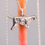 Back view of the vintage mid-century sterling silver golden retriever charm on a silver cable chain, showing the original sculptural relief detail of the feathered coat, against a soft gray background with an orange prop.