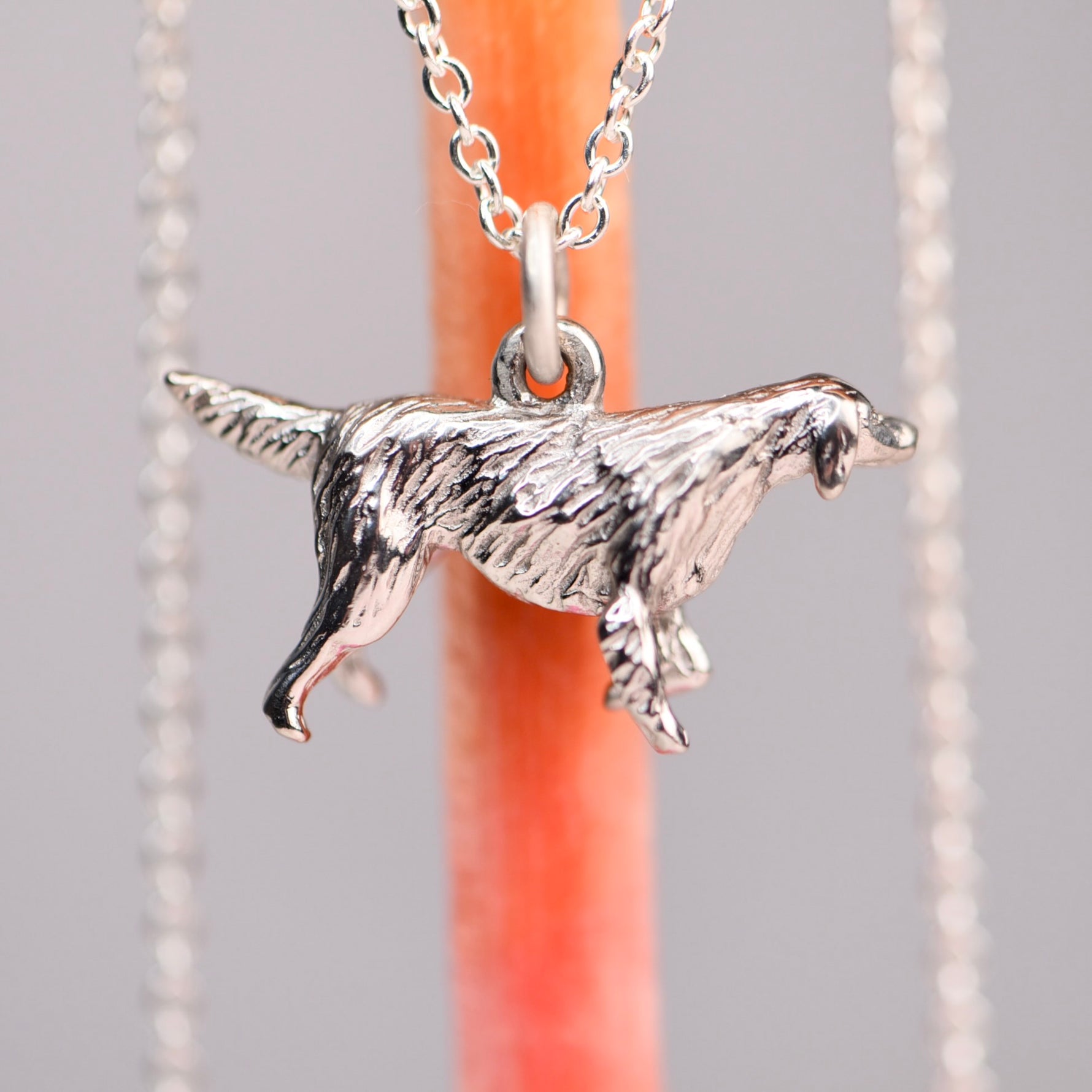 Back view of the vintage mid-century sterling silver golden retriever charm on a silver cable chain, showing the original sculptural relief detail of the feathered coat, against a soft gray background with an orange prop.