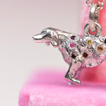 Close-up of a vintage mid-century sterling silver golden retriever charm with hand engraved star settings flush set with pink, purple, yellow, orange, red, and brown sapphires and colored diamonds against a pink background.