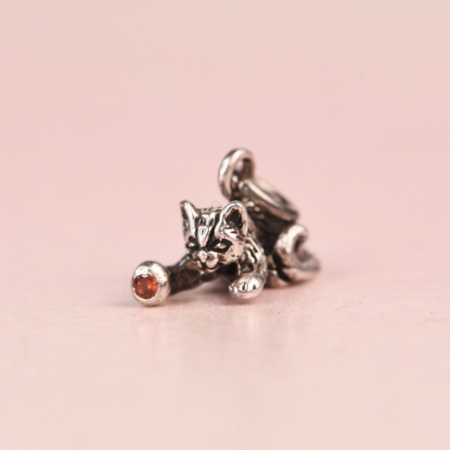 Close up of a vintage sterling silver Jezlaine kitten charm playing with a ball set with an orange diamond, on a pink background
