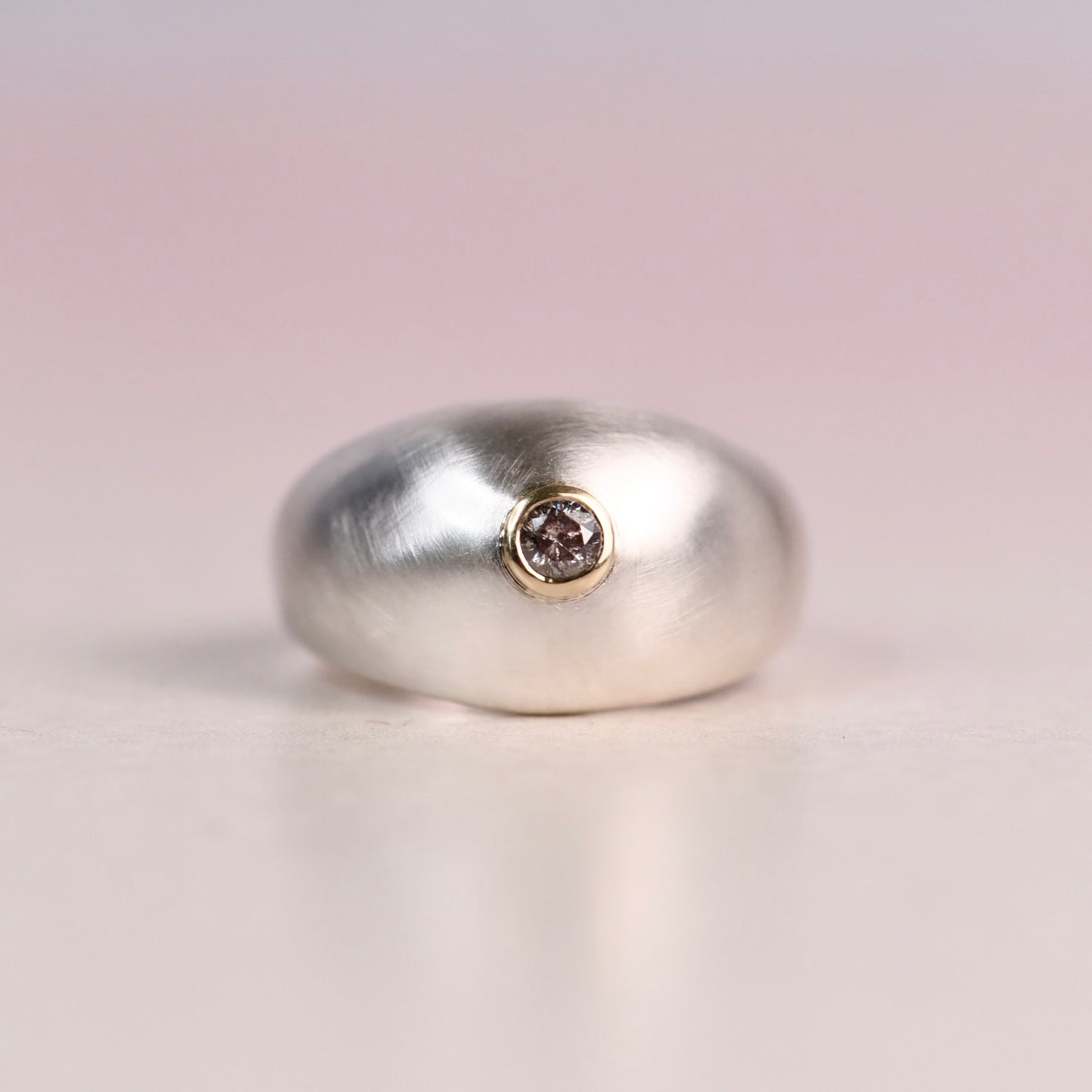 Front view of a brushed sterling silver dome ring with a 14k yellow gold bezel set with a faceted salt and pepper diamond, displayed on a white surface against a soft blush pink background.