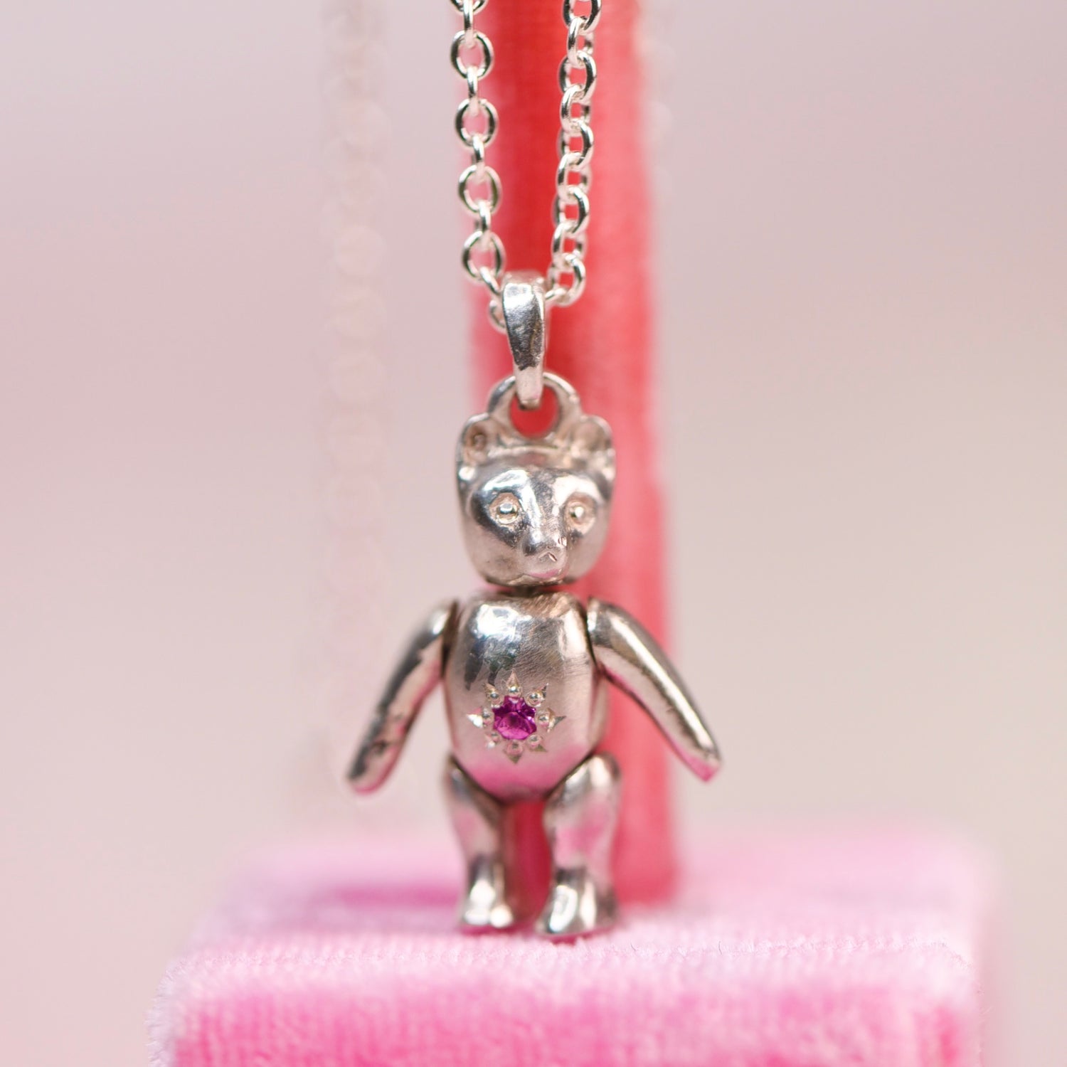 Vintage sterling silver articulating teddy bear charm necklace sitting on a pink velvet display, featuring a hand engraved star-set pink sapphire belly charm