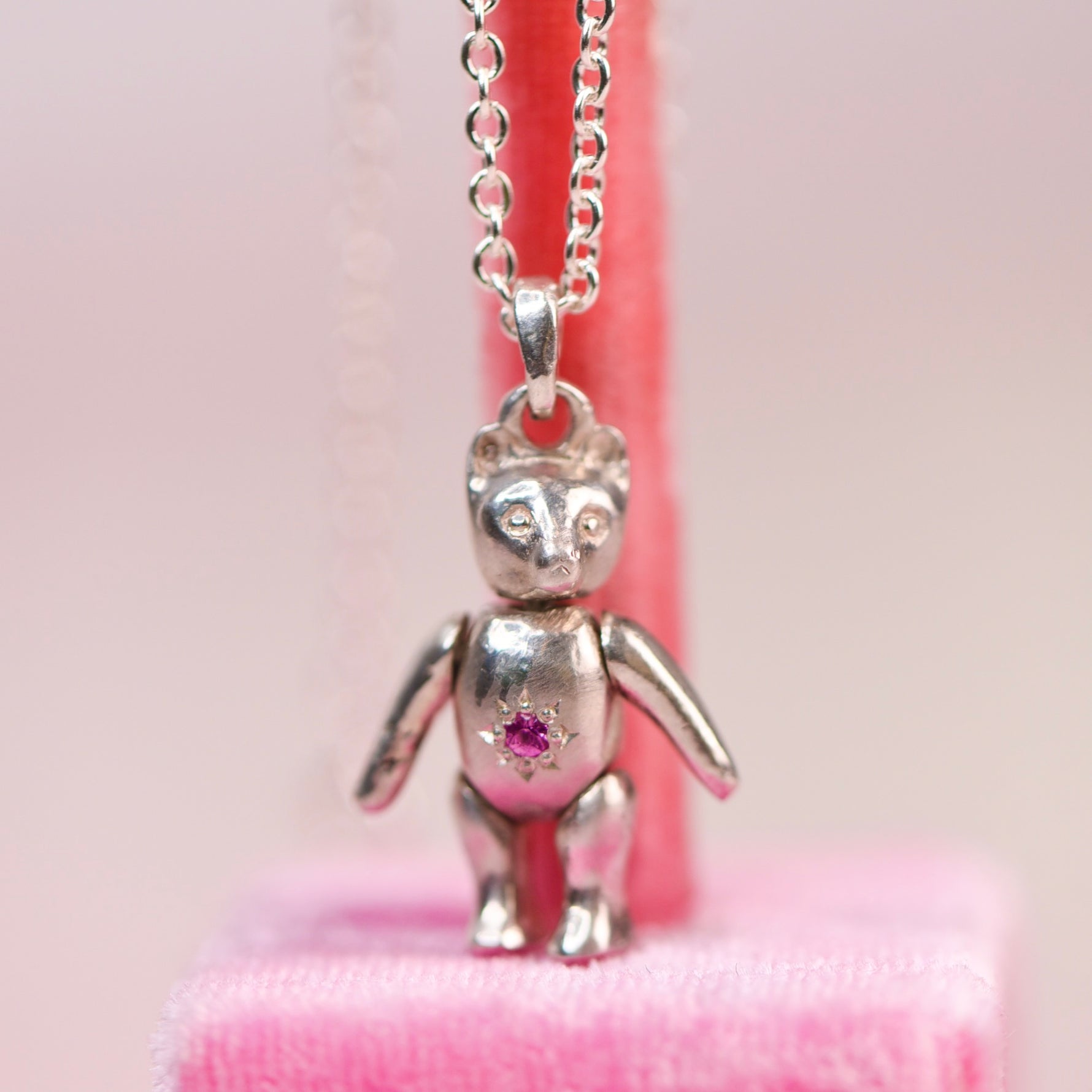 Vintage sterling silver articulating teddy bear charm necklace sitting on a pink velvet display, featuring a hand engraved star-set pink sapphire belly charm