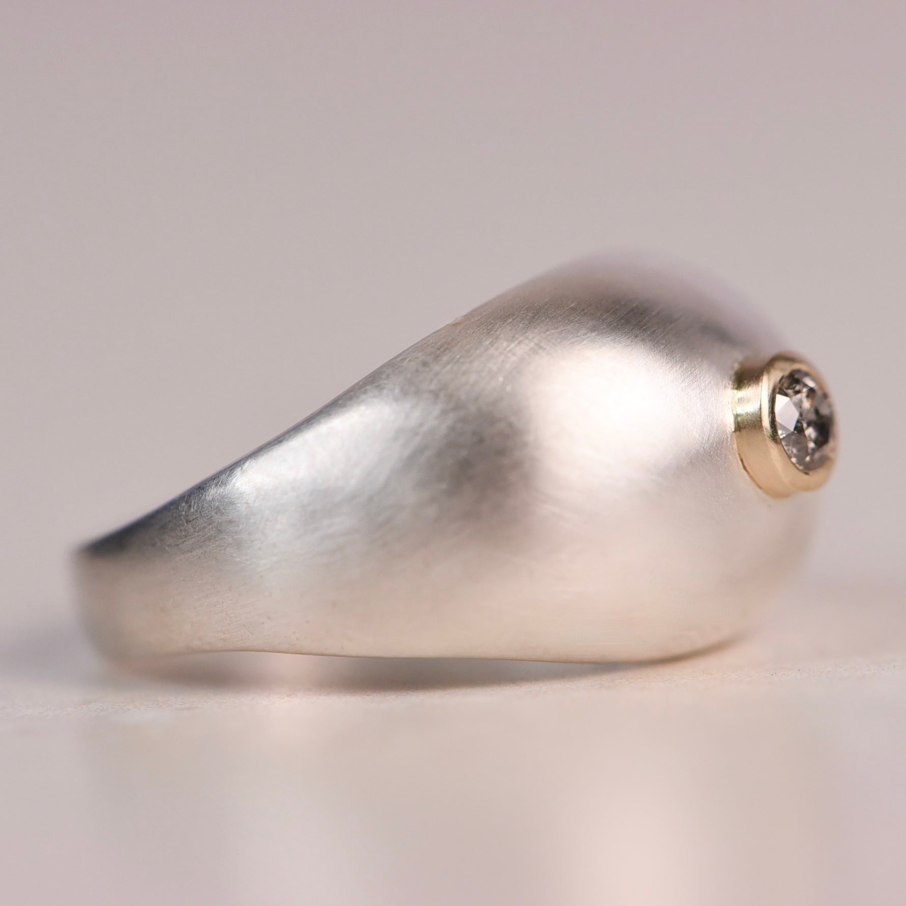 Side profile of a brushed sterling silver dome ring with a 14k yellow gold bezel set with a faceted salt and pepper diamond, displayed on a white surface against a soft grey background.