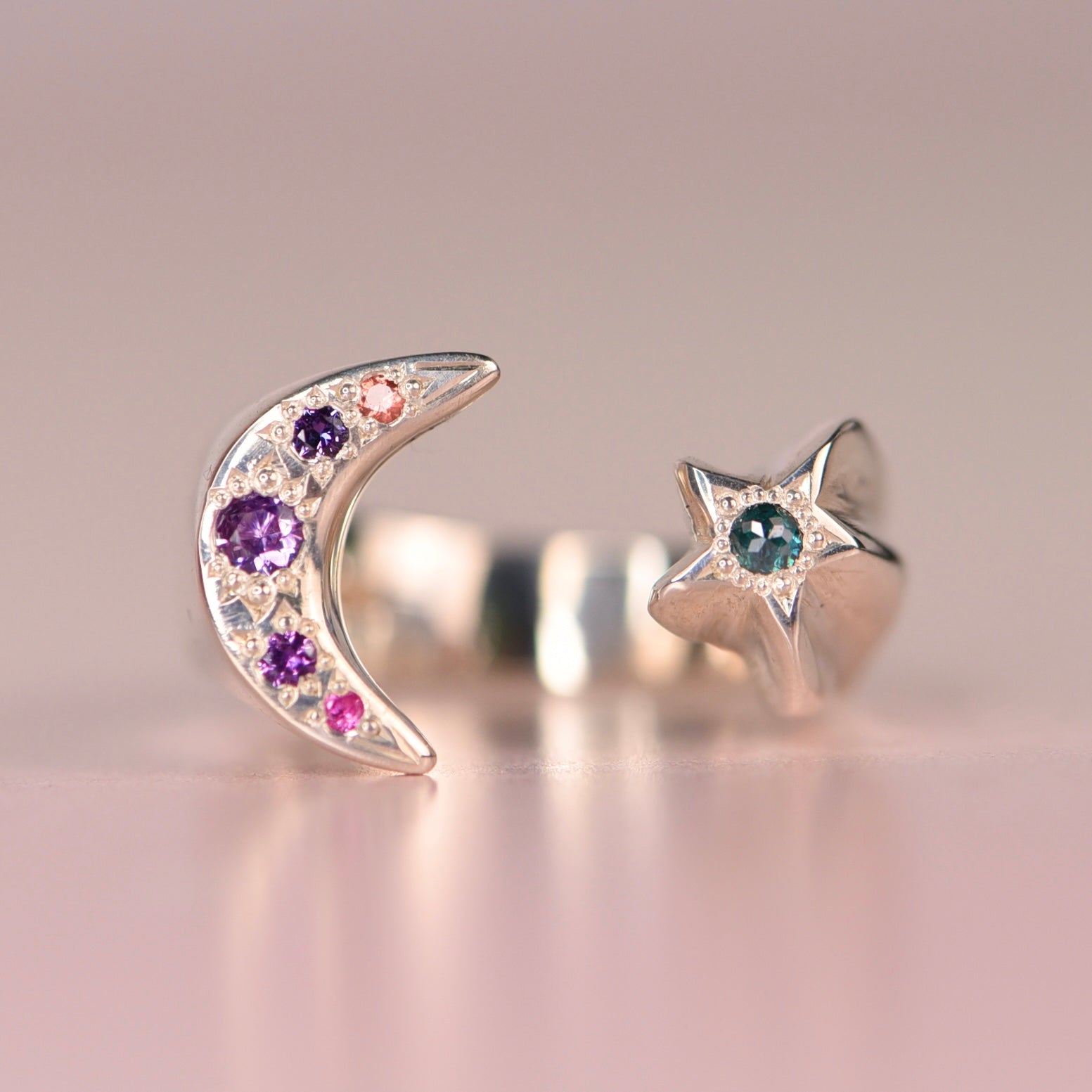 Vintage sterling silver open moon and star ring with hand engraved purple and pink sapphires on the crescent moon and a green rose-cut diamond on the star, displayed on a pink surface by Corkie Bolton Jewelry