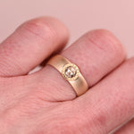 Close-up of a wide brushed 14k yellow gold ring with a hand-engraved 18k gold bezel set with a salt and pepper rose cut diamond, worn on a hand against a soft blush pink background.