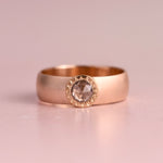 A wide polished yellow gold band ring with a smooth rounded profile, displayed upright on a soft blush pink reflective surface against a light grey background.