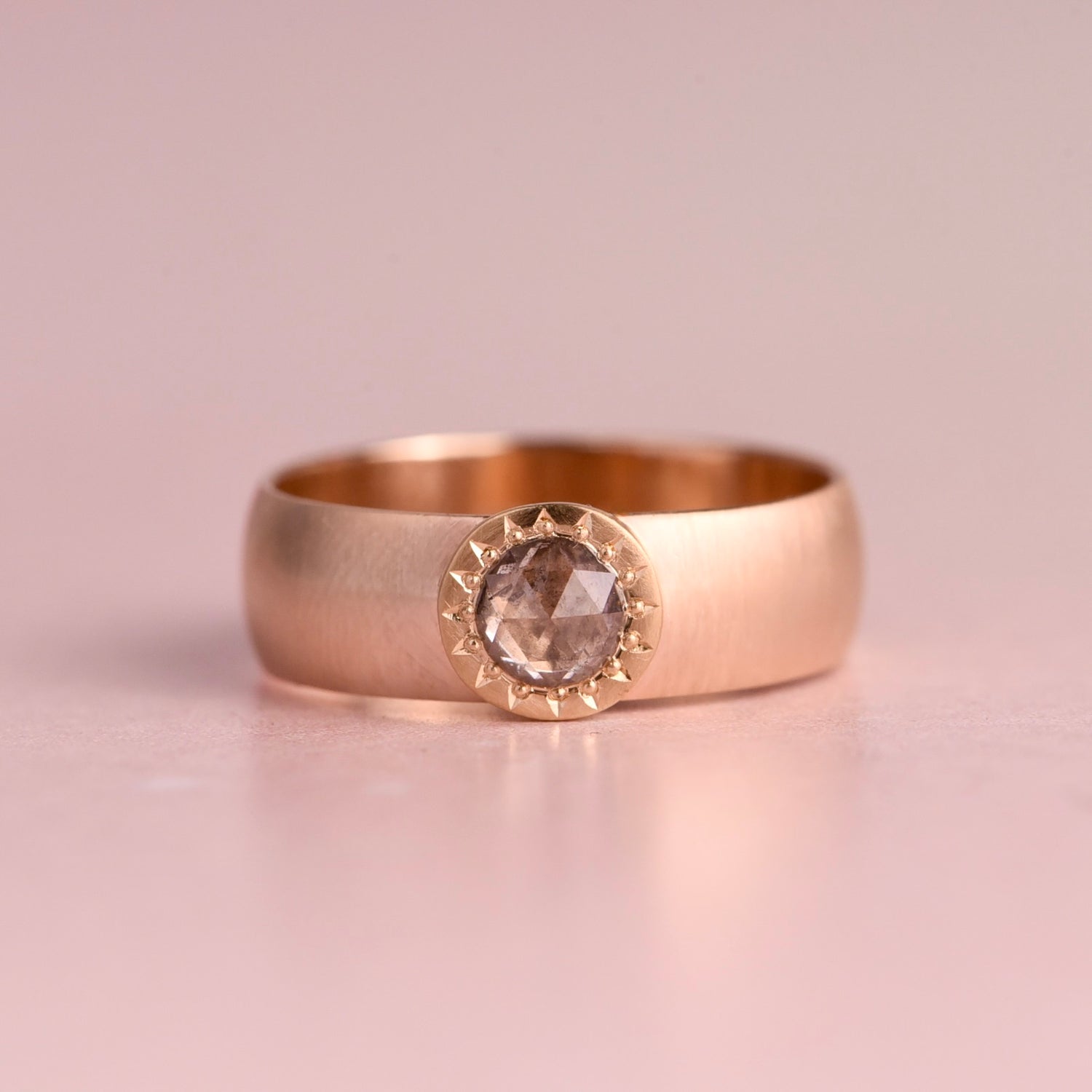 A wide polished yellow gold band ring with a smooth rounded profile, displayed upright on a soft blush pink reflective surface against a light grey background.
