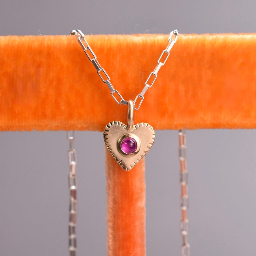 14k Gold heart-shaped pendant with a pink ruby on a silver chain, displayed on an orange stand.