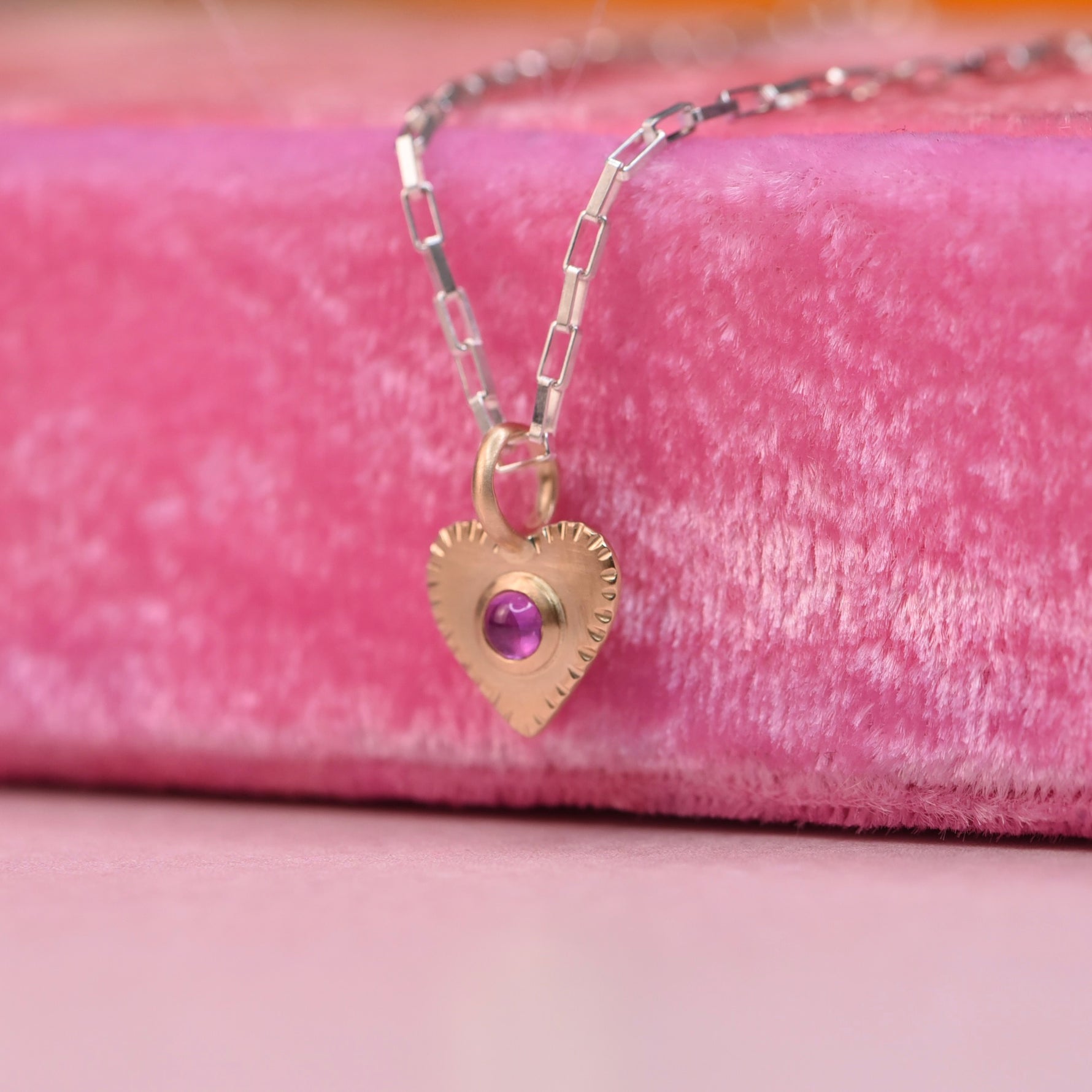 Engraving details side angle of 14k Gold heart-shaped pendant with a pink ruby on a silver chain, displayed on a neon pink velvet display